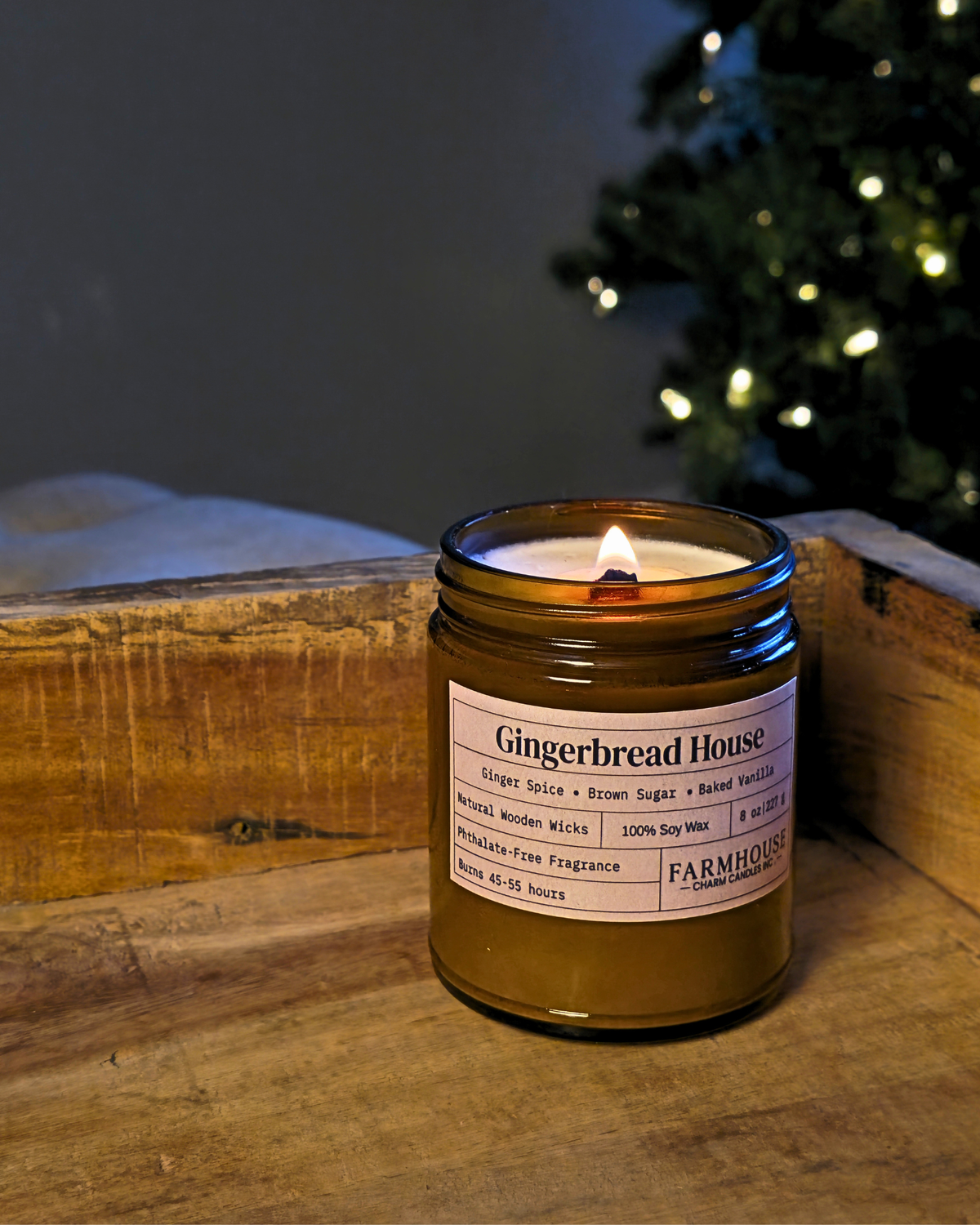 A lit amber glass jar candle labeled 'Gingerbread House' by Farmhouse Charm Candles placed on a rustic wooden surface with a blurred Christmas tree in the background.