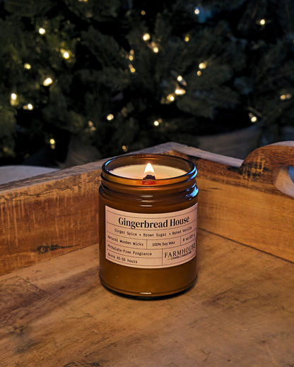 A lit amber glass jar candle labeled 'Gingerbread House' by Farmhouse Charm Candles placed on a rustic wooden surface with a blurred Christmas tree and warm string lights in the background.
