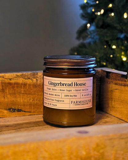 A Farmhouse Charm Candles. Gingerbread House scented wax candle in an amber glass jar with a dark metal lid, resting on a rustic wooden crate with a blurred Christmas tree in the background.