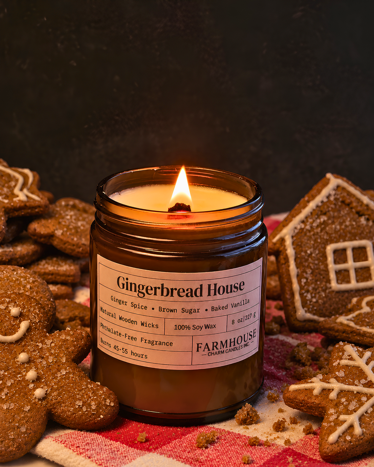 A lit amber glass jar candle labeled 'Gingerbread House' by Farmhouse Charm Candles styled with gingerbread cookies and a house-shaped cookie on a red-and-white checkered cloth against a dark background.
