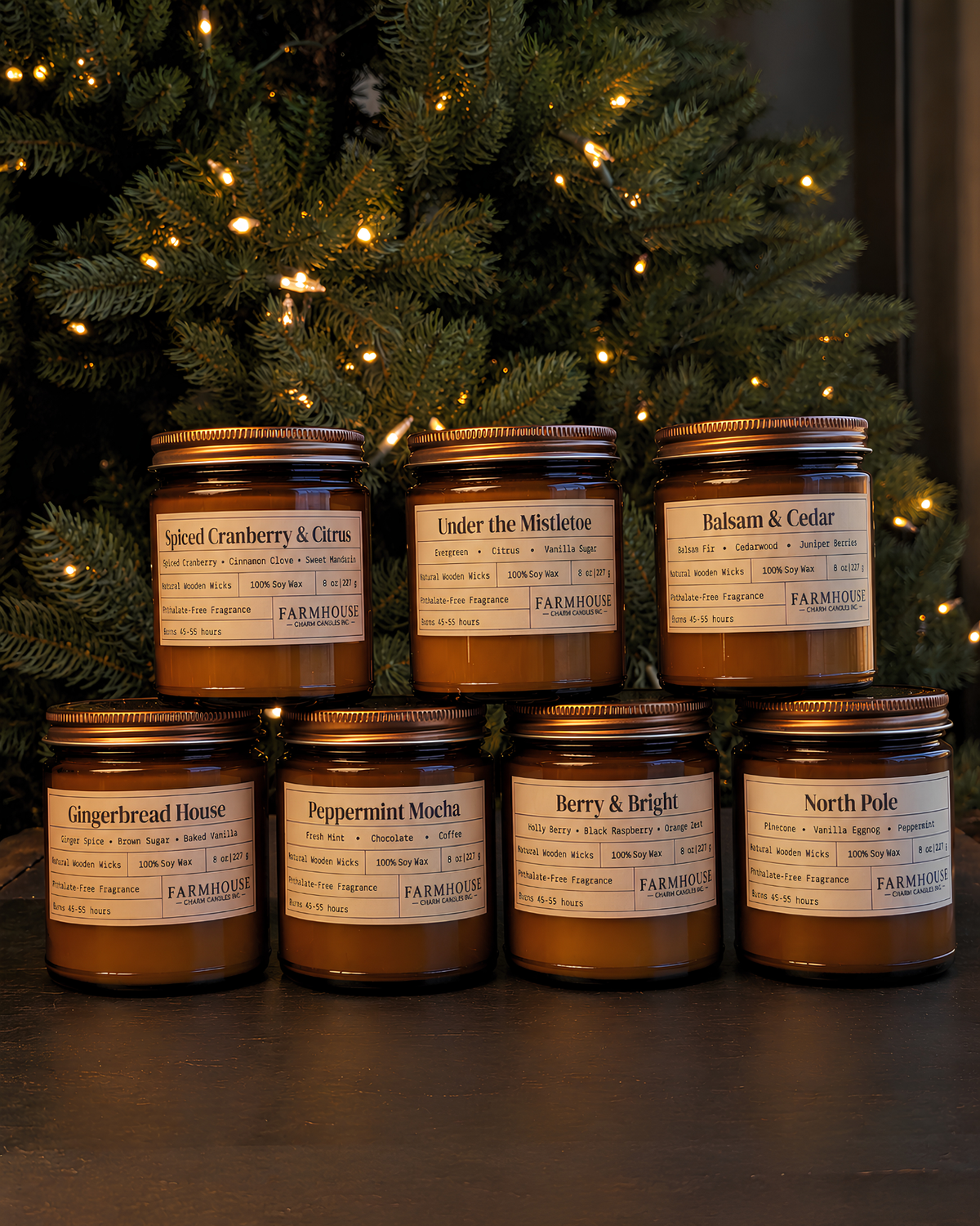 7 amber glass jar candles with cream labels and gold lids are arranged in two rows in front of a lit Christmas tree. The labels show different scent names including Spiced Cranberry & Carom, Under the Mistletoe, Balsam & Cedar, Gingerbread House, Peppermint Mocha, Berry & Bright, and North Pole. Home for Christmas Candle Bundle by Farmhouse Charm Candles