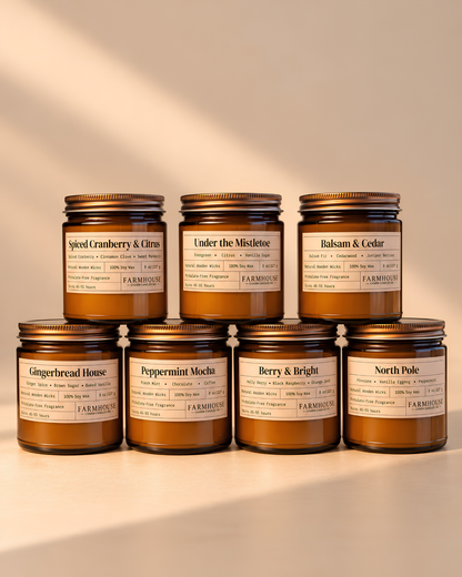 7 amber glass jar candles with gold lids and cream labels arranged in two rows on a warm beige surface. The labels show different scent names including Spiced Cranberry & Citrus, Under the Mistletoe, Balsam & Cedar, Gingerbread House, Peppermint Mocha, Berry & Bright, and North Pole. Home for Christmas Candle Bundle by Farmhouse Charm Candles