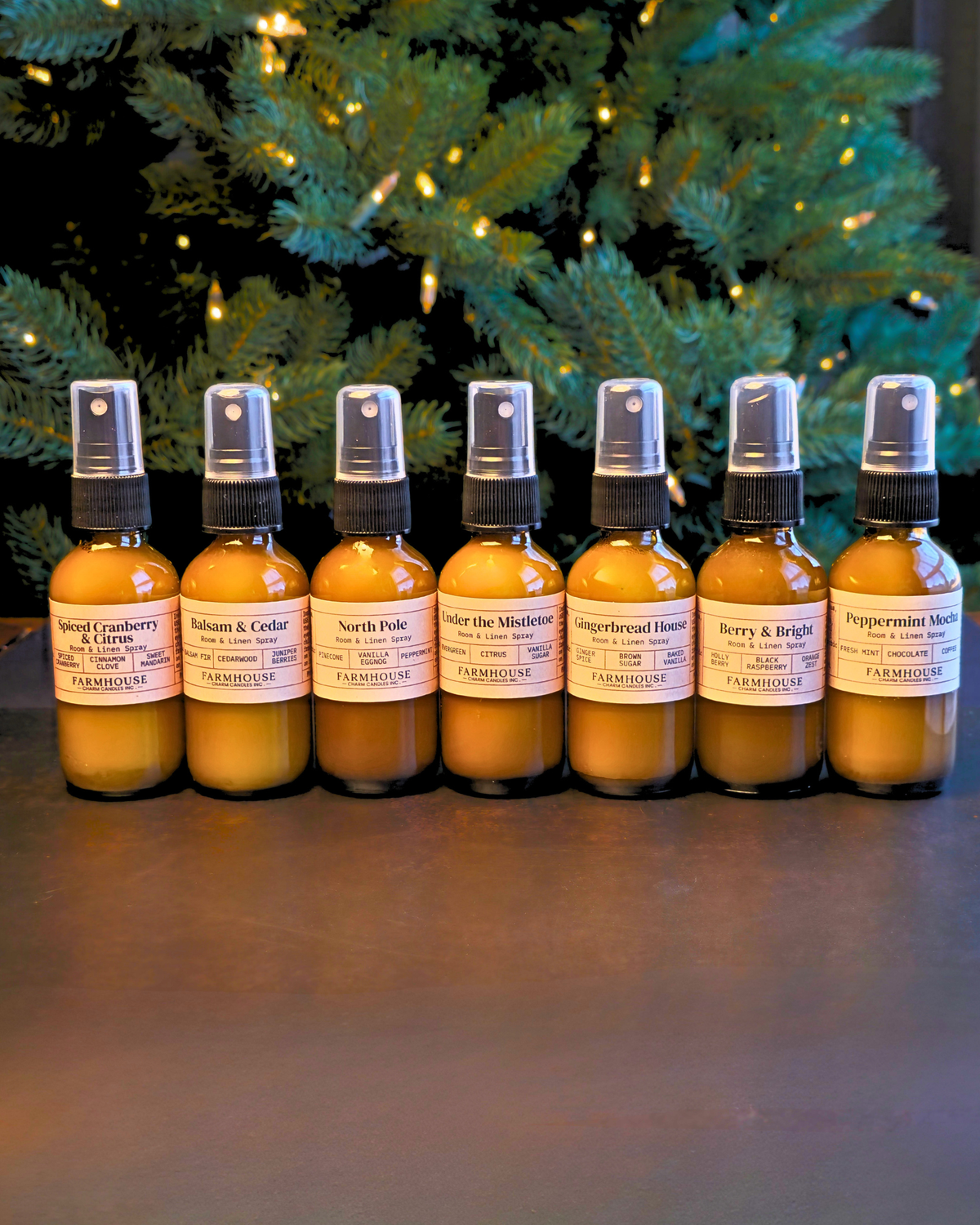 Seven amber glass spray bottles with white labels arranged in a row on a dark surface, with a blurred Christmas tree in the background. Home For Christmas Room & Linen Spray by Farmhouse Charm Candles