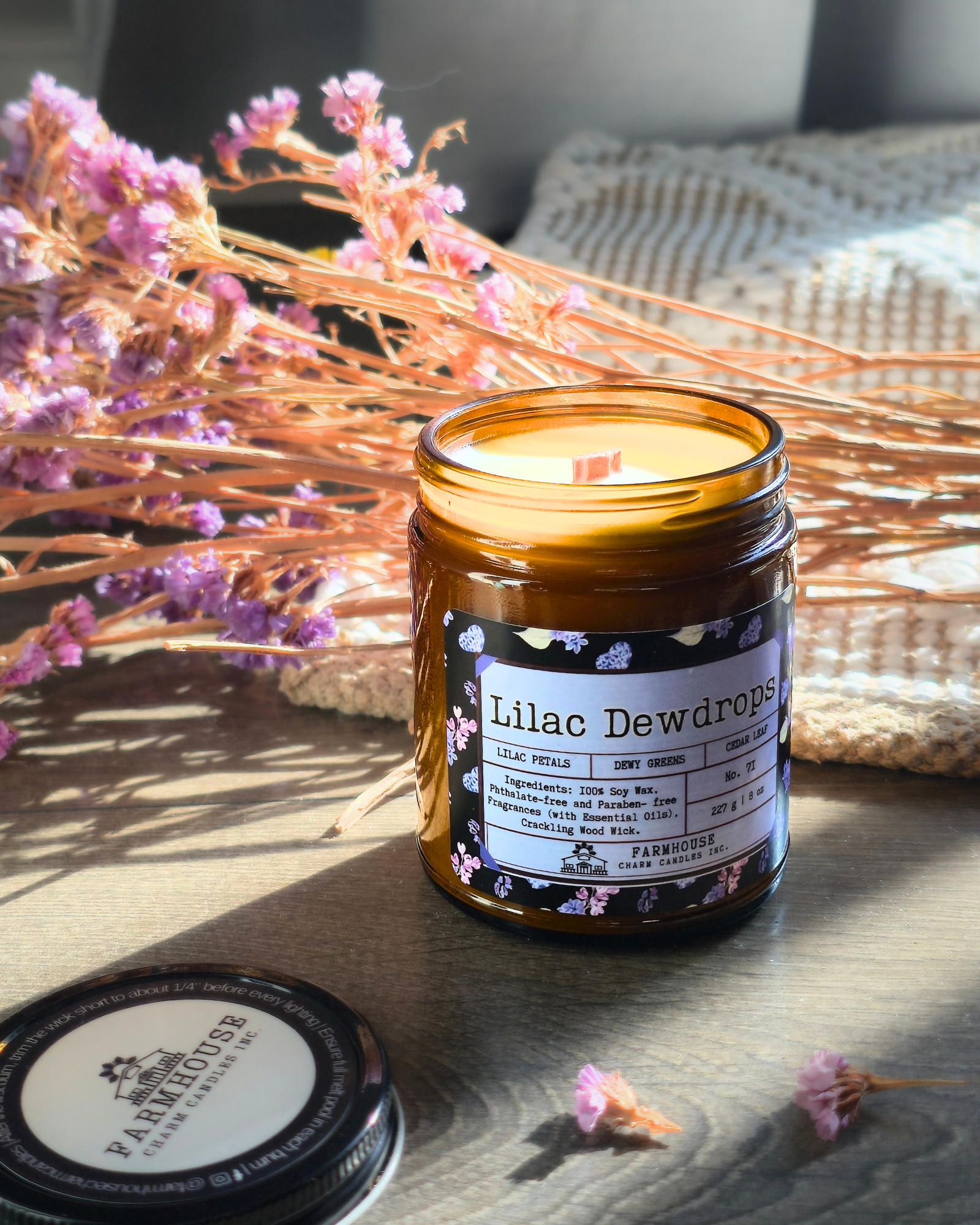 Candle in an amber jar labeled 'Lilac Dewdrops' with dried flowers and a woven basket in the background. Farmhouse Charm Candles