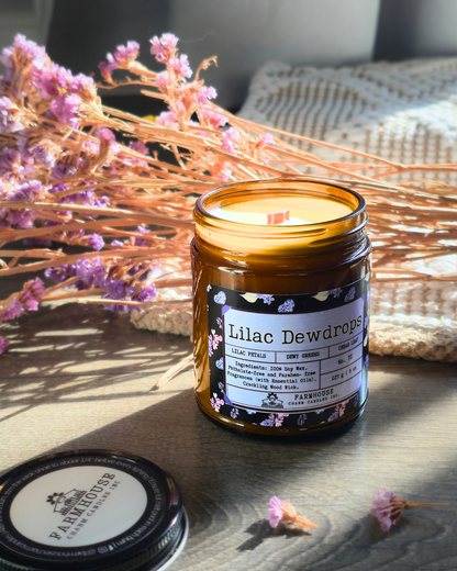 Candle in an amber jar labeled 'Lilac Dewdrops' with dried flowers and a woven basket in the background. Farmhouse Charm Candles