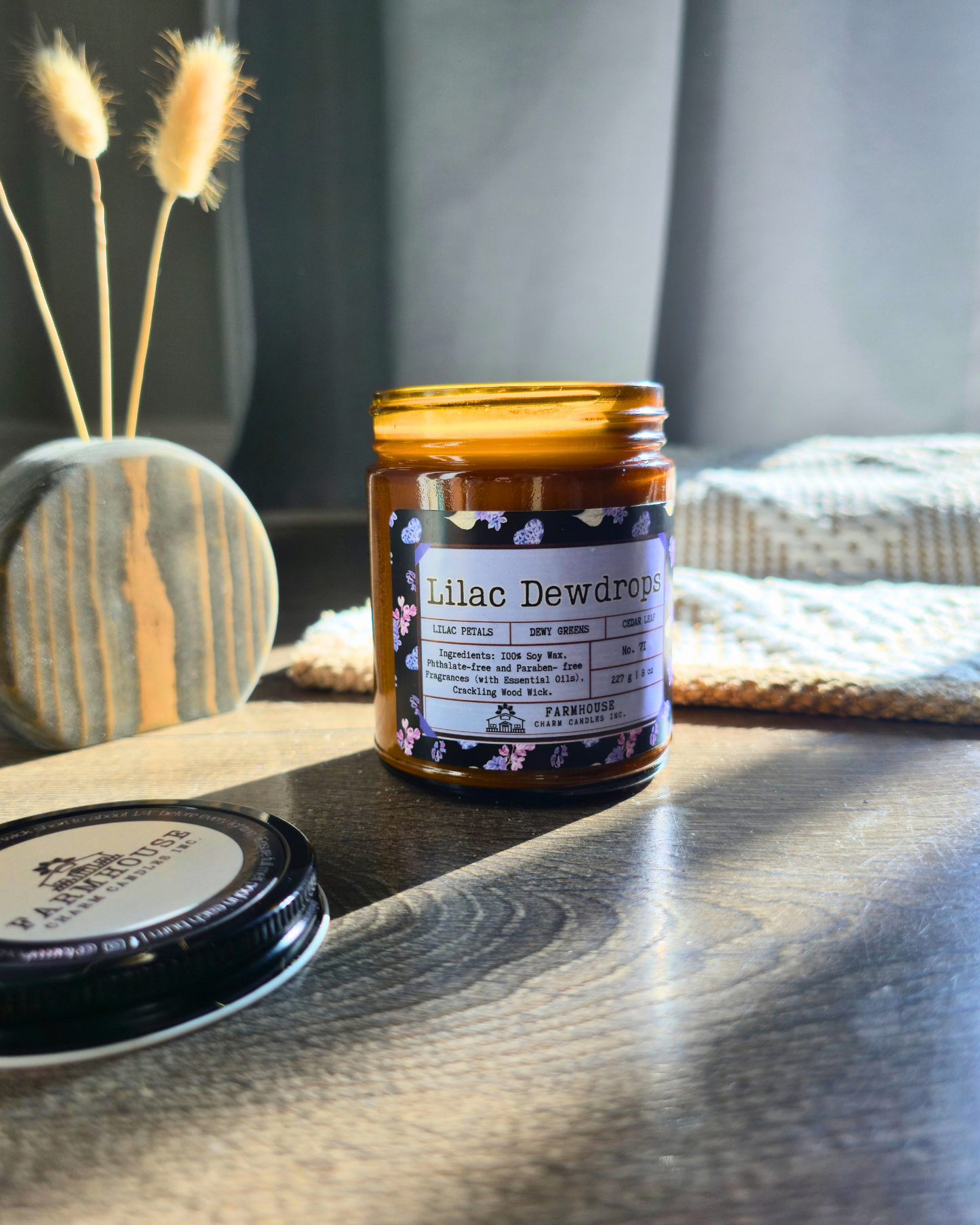 Jar of 'Lilac Dewdrops' candle on a wooden surface with a cozy background. Farmhouse Charm Candles