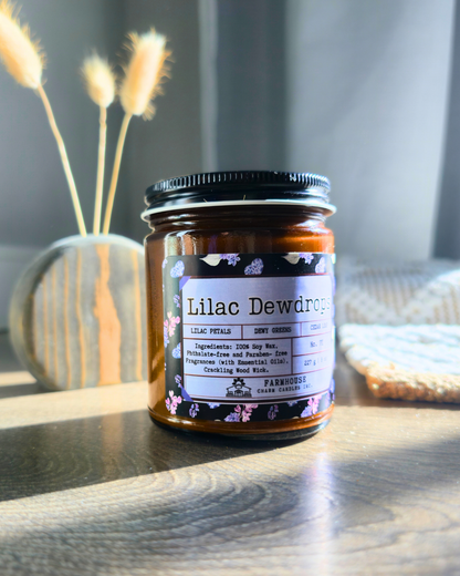 Jar of 'Lilac Dewdrops' candle on a wooden surface with a blurred background. Farmhouse Charm Candles