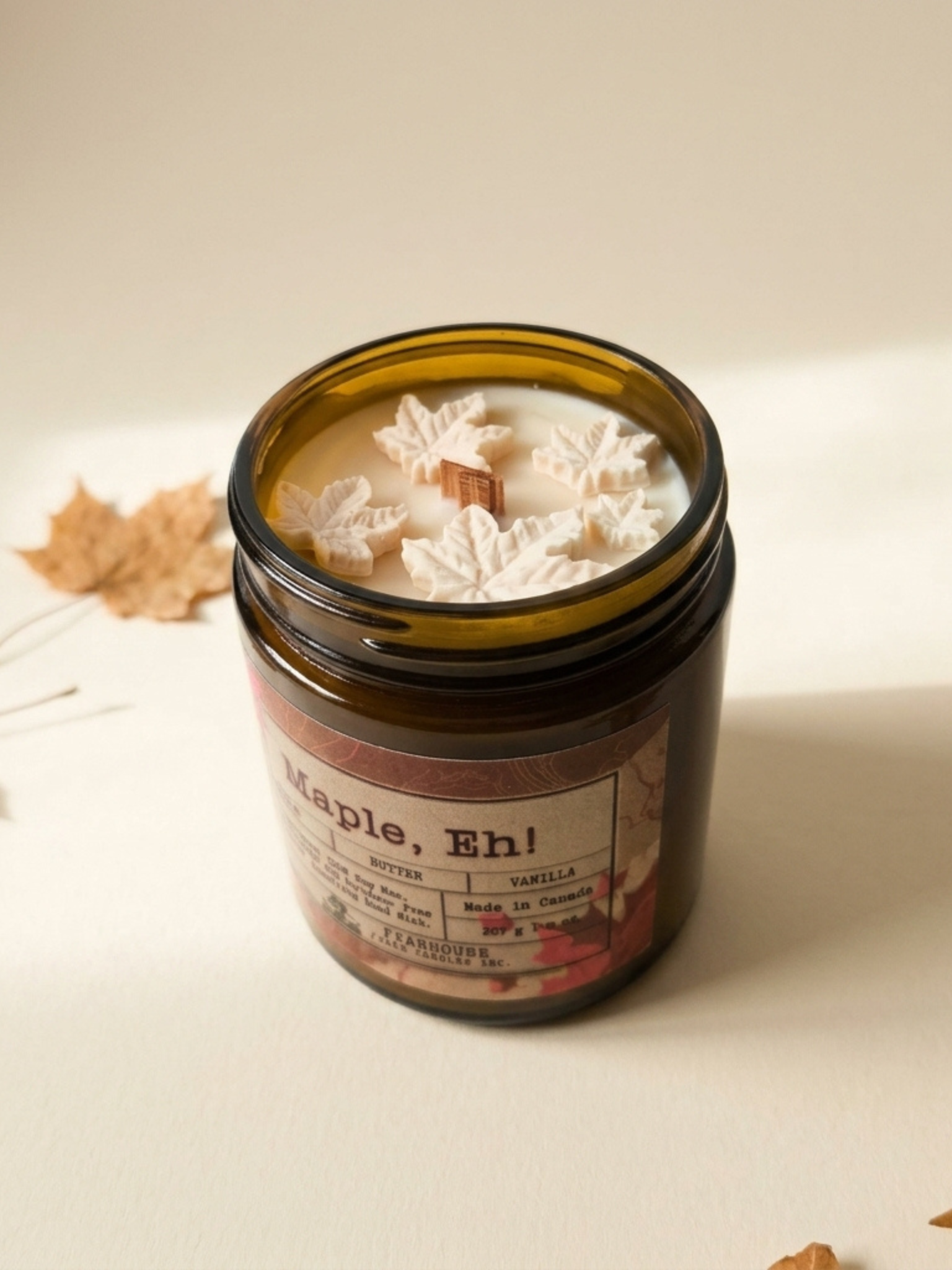 Candle in a glass jar labeled 'Maple, Eh!' by Farmhouse Charm Candles on a light background