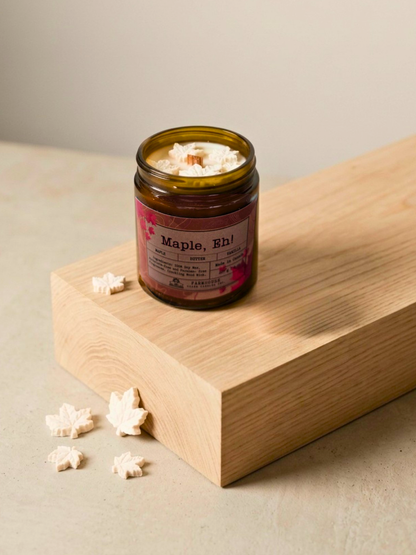 Jar of candle labeled 'Maple, Eh' by Farmhouse Charm Candles on a wooden surface with popcorn around