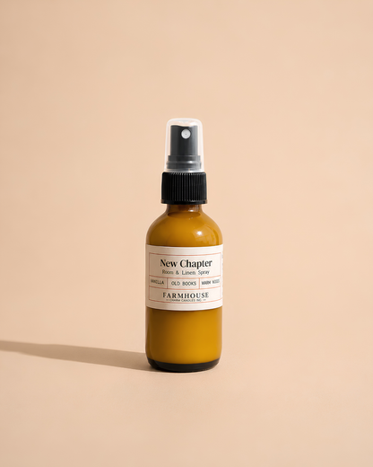 A small amber glass spray bottle with a black pump and a white label reading 'New Chapter' by Farmhouse Charm Candles photographed on a warm beige studio background.