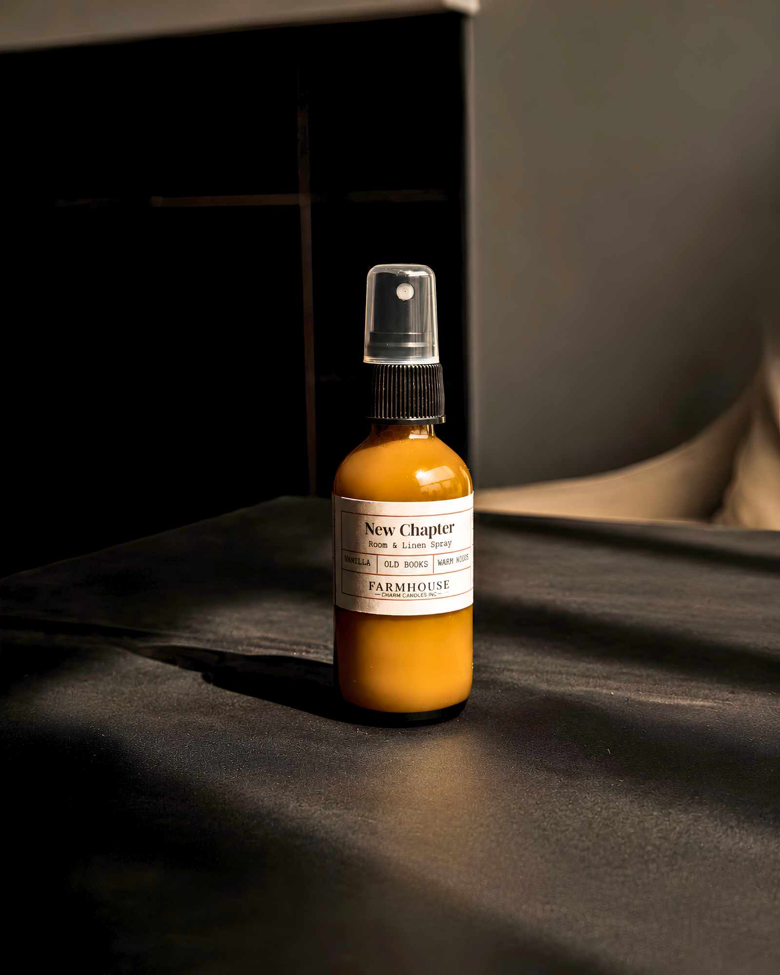 A small amber spray bottle with a white label reading 'New Chapter' by Farmhouse Charm Candles stands on a dark tabletop in moody indoor lighting.