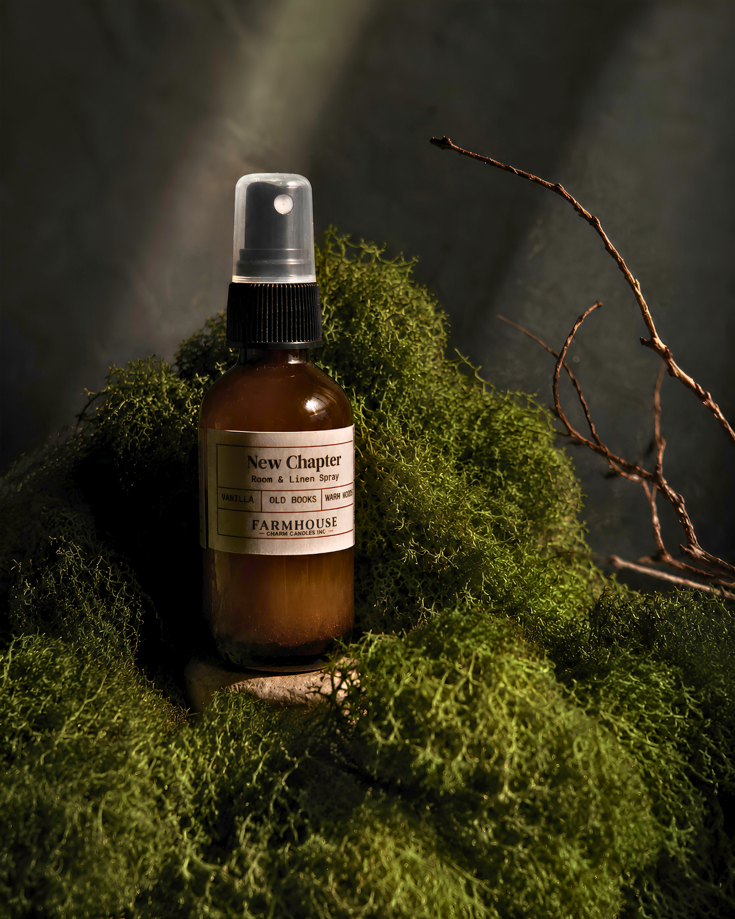 An amber glass spray bottle with a black sprayer and a cream label reading 'New Chapter' by Farmhouse Charm Candles, placed among green moss on a dark textured background with a dried branch in the upper right.