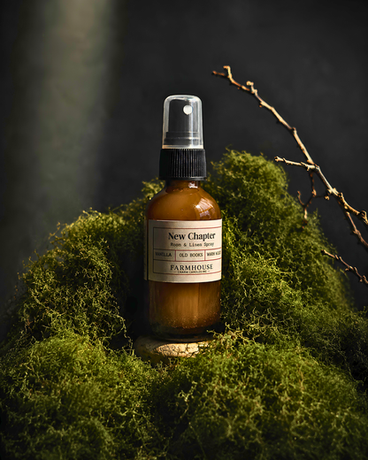 An amber glass spray bottle labeled 'New Chapter by Farmhouse Charm Candles stands on a stone among dense green moss against a dark, moody background with a bare branch reaching in from the upper right.