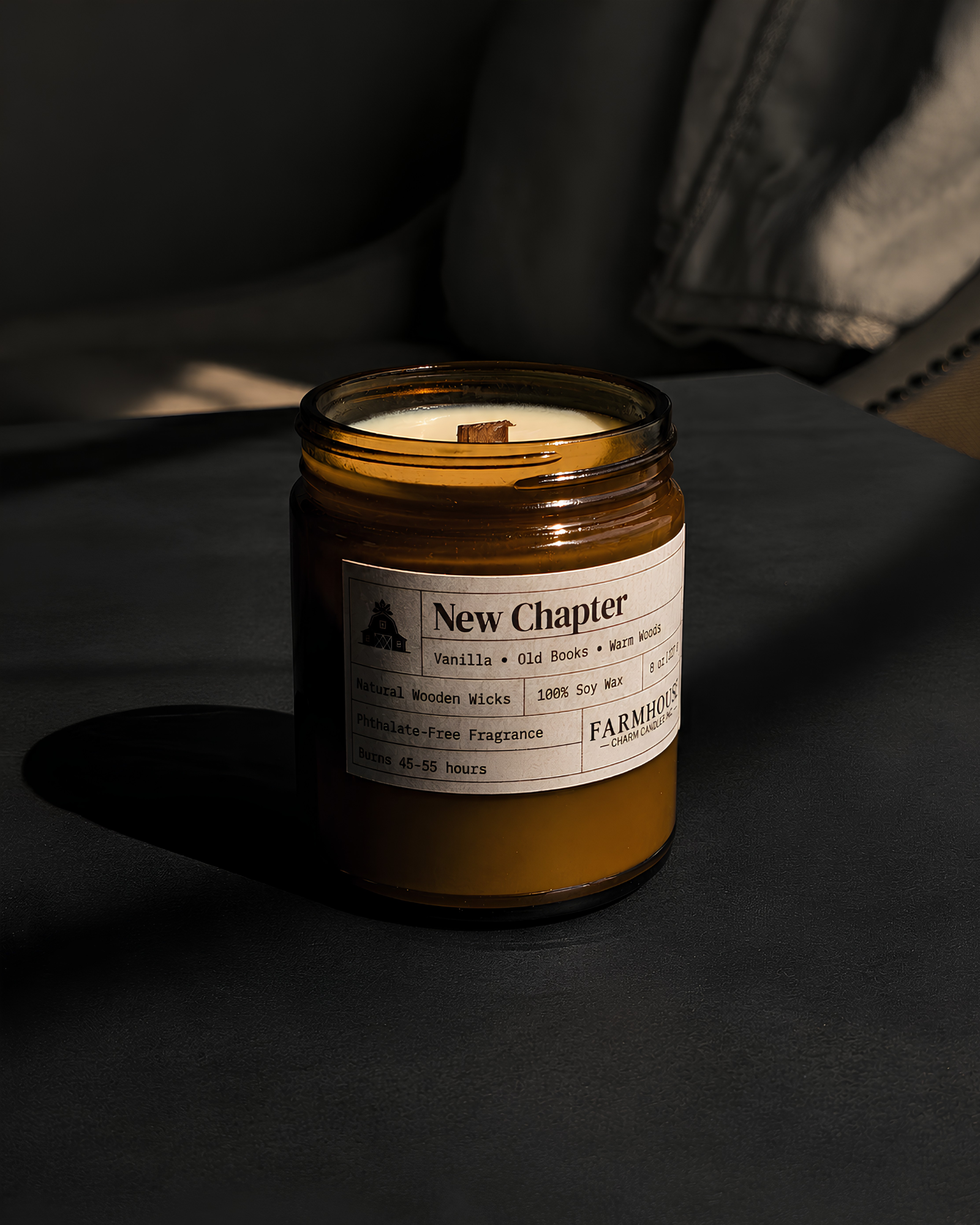 An amber glass jar candle with a white label reading 'New Chapter'  by Farmhouse Charm Candles on a dark tabletop, with a chair and strong sunlight in the background.
