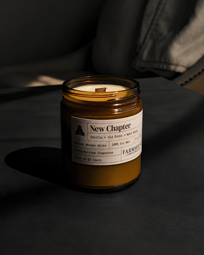 An amber glass jar candle with a white label reading 'New Chapter'  by Farmhouse Charm Candles on a dark tabletop, with a chair and strong sunlight in the background.