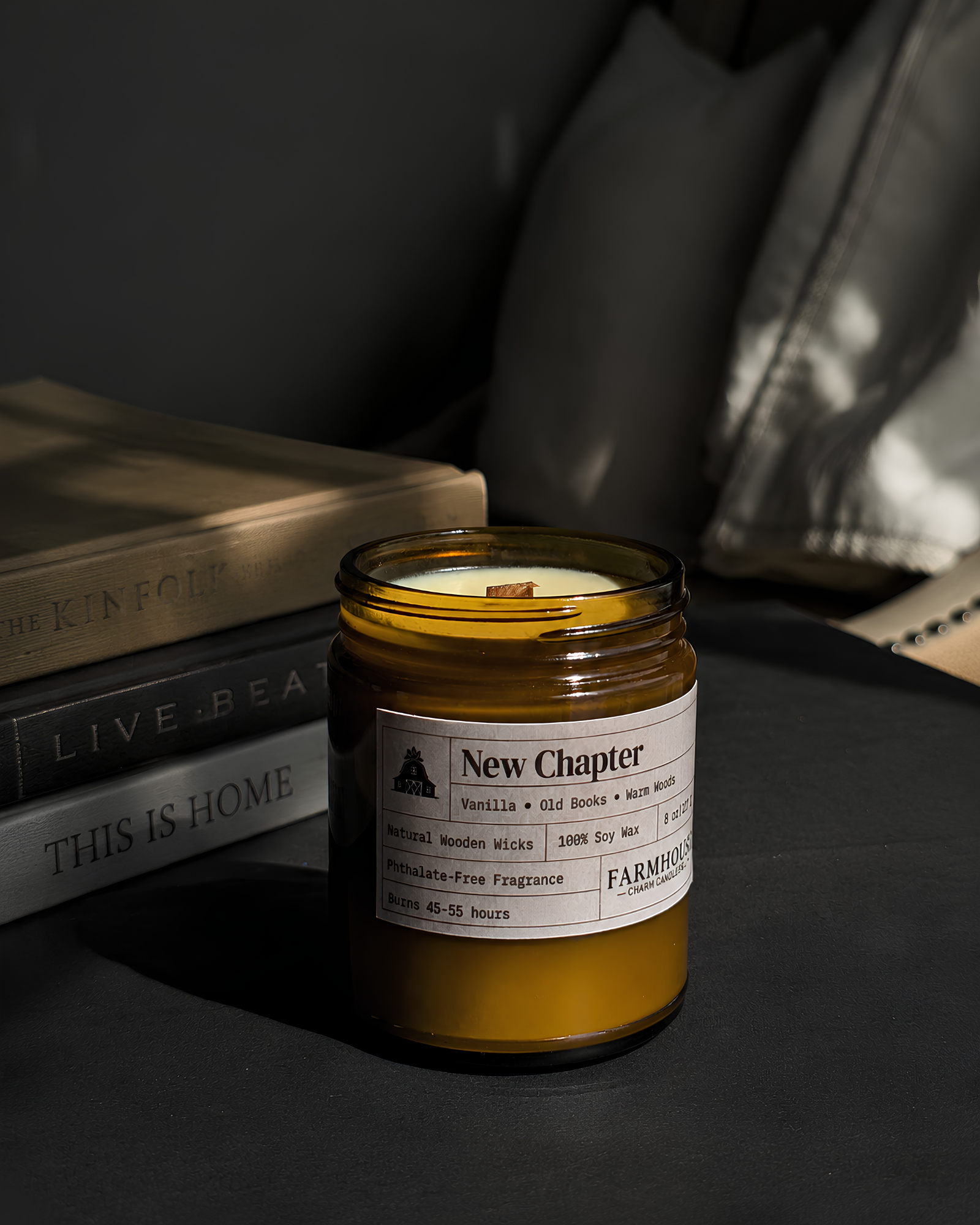 A New Chapter candle in an amber glass jar sits on a dark surface next to stacked books, with a soft gray pillow in the background. The candle wax is a warm golden yellow, and the label reads 'New Chapter' by Farmhouse Charm Candles