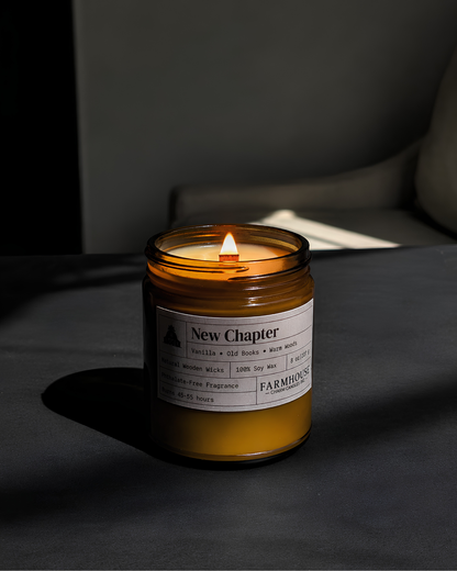 A lit amber glass jar candle on a dark tabletop in a moody room setting. The candle label reads 'New Chapter' by Farmhouse Charm Candles, with additional scent and ingredient text visible.