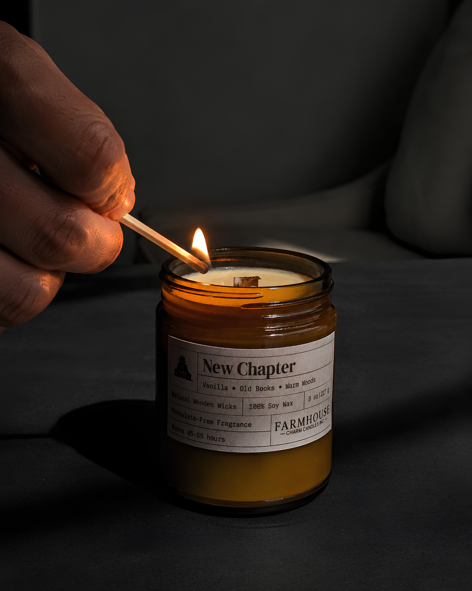 A hand holding a lit match to ignite a New Chapter Farmhouse scented candle in an amber glass jar, set against a dark, moody background.