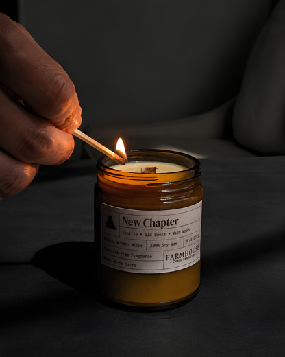 A hand holding a lit match to ignite a New Chapter Farmhouse scented candle in an amber glass jar, set against a dark, moody background.