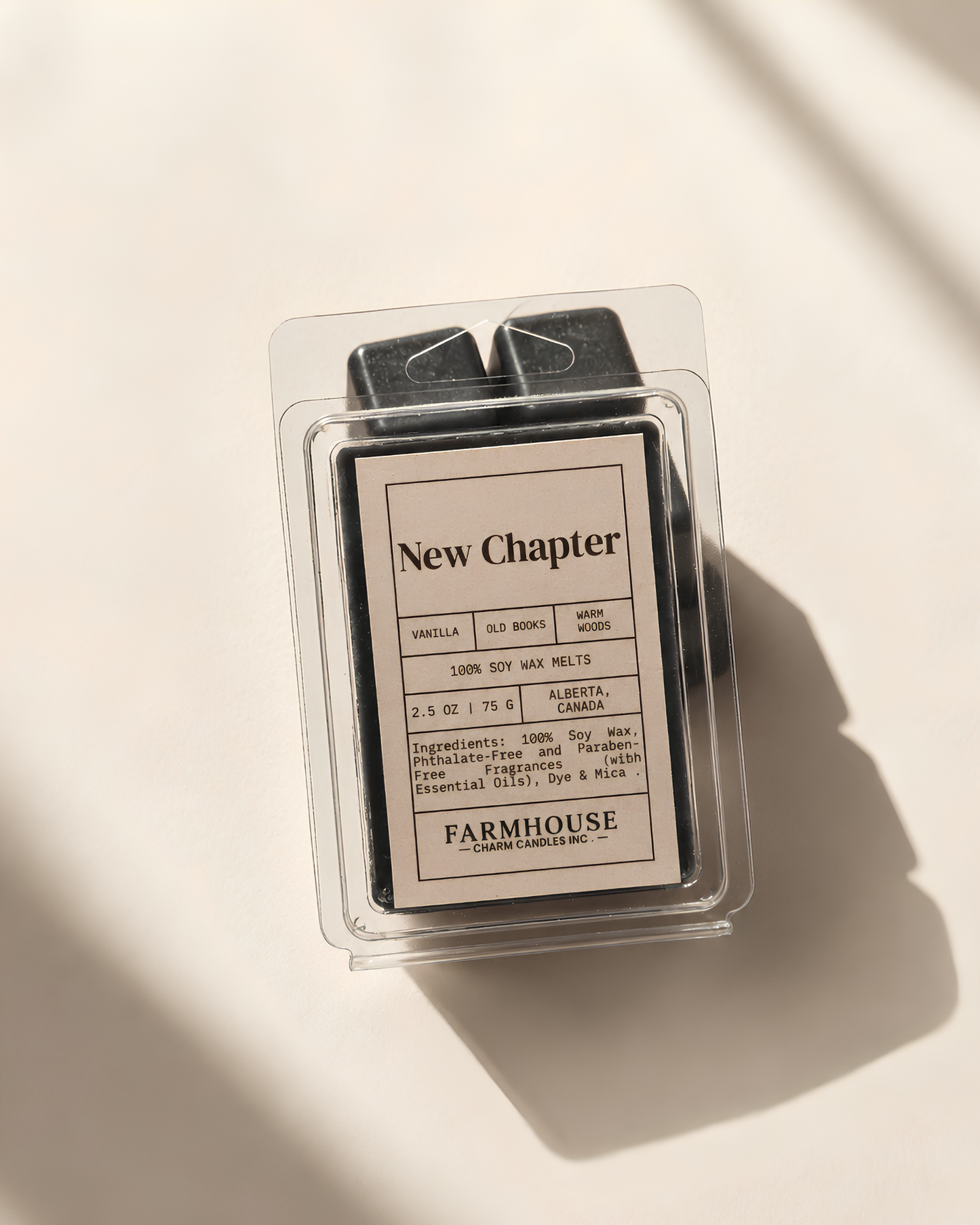 A wax melt package from Farmhouse Charm Candles. under the New Chapter scent, shown in a clear plastic clamshell with a cream-colored label and black wax melts visible inside, photographed on a warm off-white surface with soft diagonal shadows.