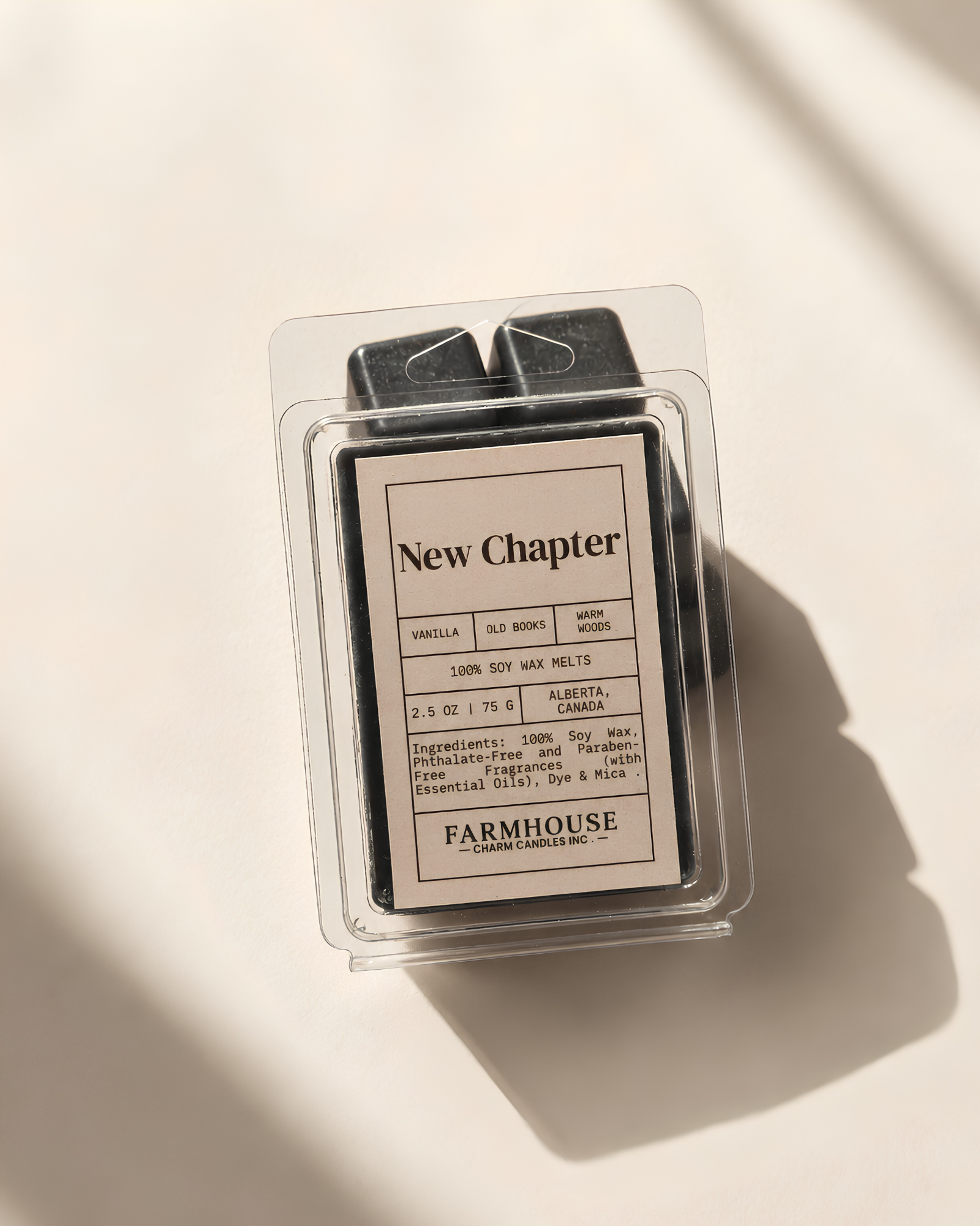A wax melt package from Farmhouse Charm Candles. under the New Chapter scent, shown in a clear plastic clamshell with a cream-colored label and black wax melts visible inside, photographed on a warm off-white surface with soft diagonal shadows.