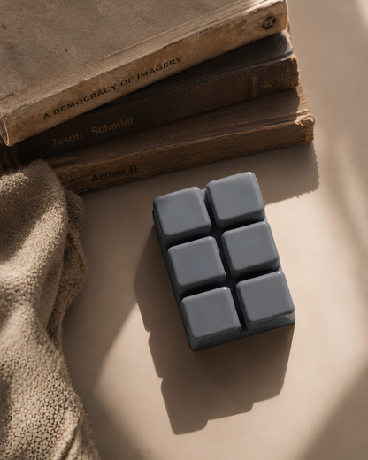A rectangular bar of dark gray soap with six square sections, styled on a beige surface next to a textured beige fabric and a stack of vintage books. Farmhouse Charm Candles