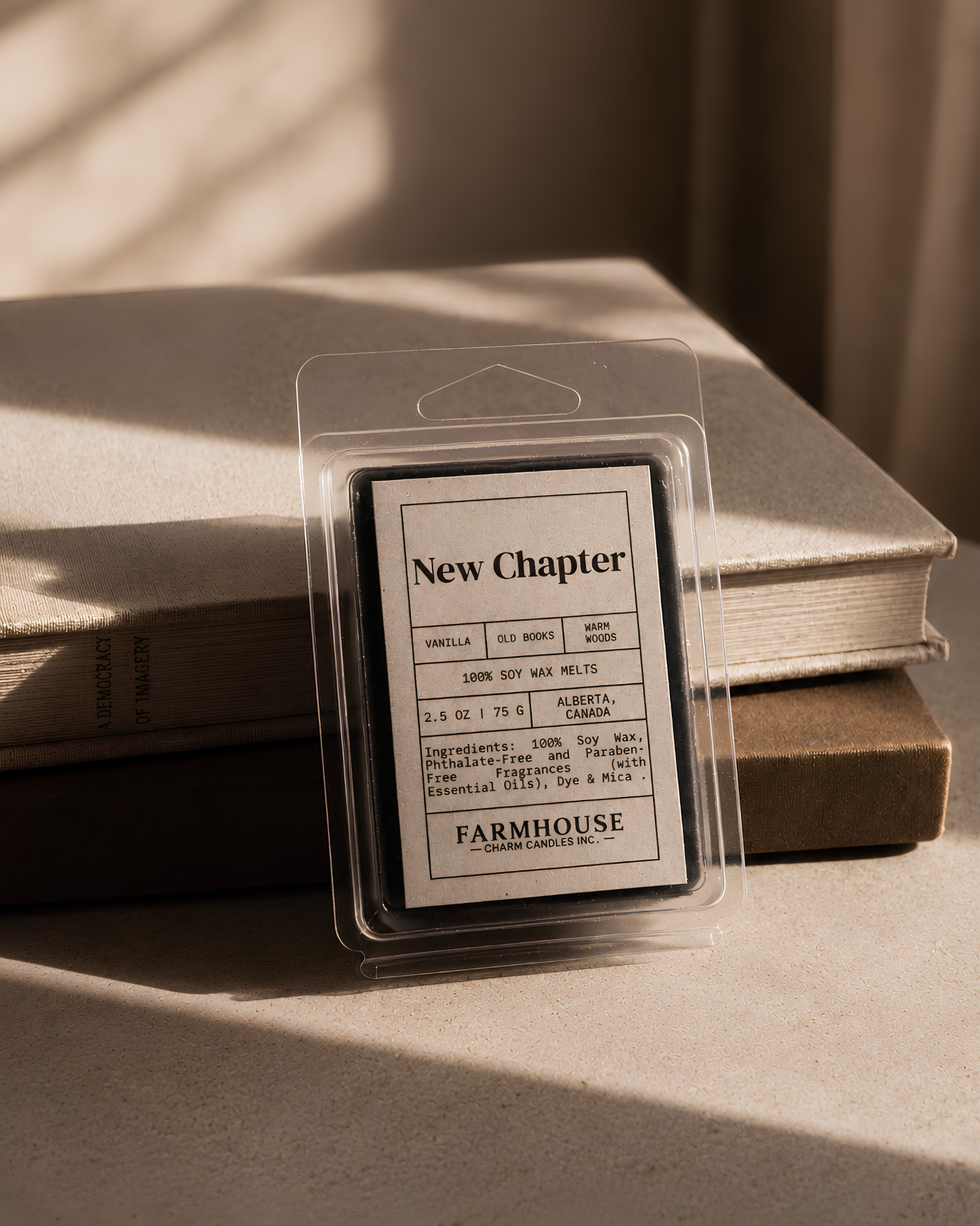 A packaged wax melt or scented wax product labeled 'New Chapter' by Farmhouse Charm Candles, stands upright in front of stacked books on a tabletop, with warm sunlight casting shadows across the scene.