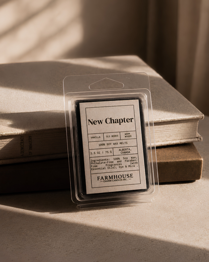 A packaged wax melt or scented wax product labeled 'New Chapter' by Farmhouse Charm Candles, stands upright in front of stacked books on a tabletop, with warm sunlight casting shadows across the scene.