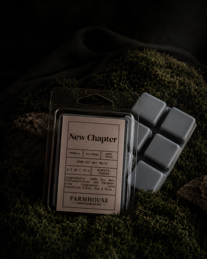 A packaged wax melt product labeled 'New Chapter' by Farmhouse Charm Candles, placed on dark mossy fabric with several gray wax cubes beside it.