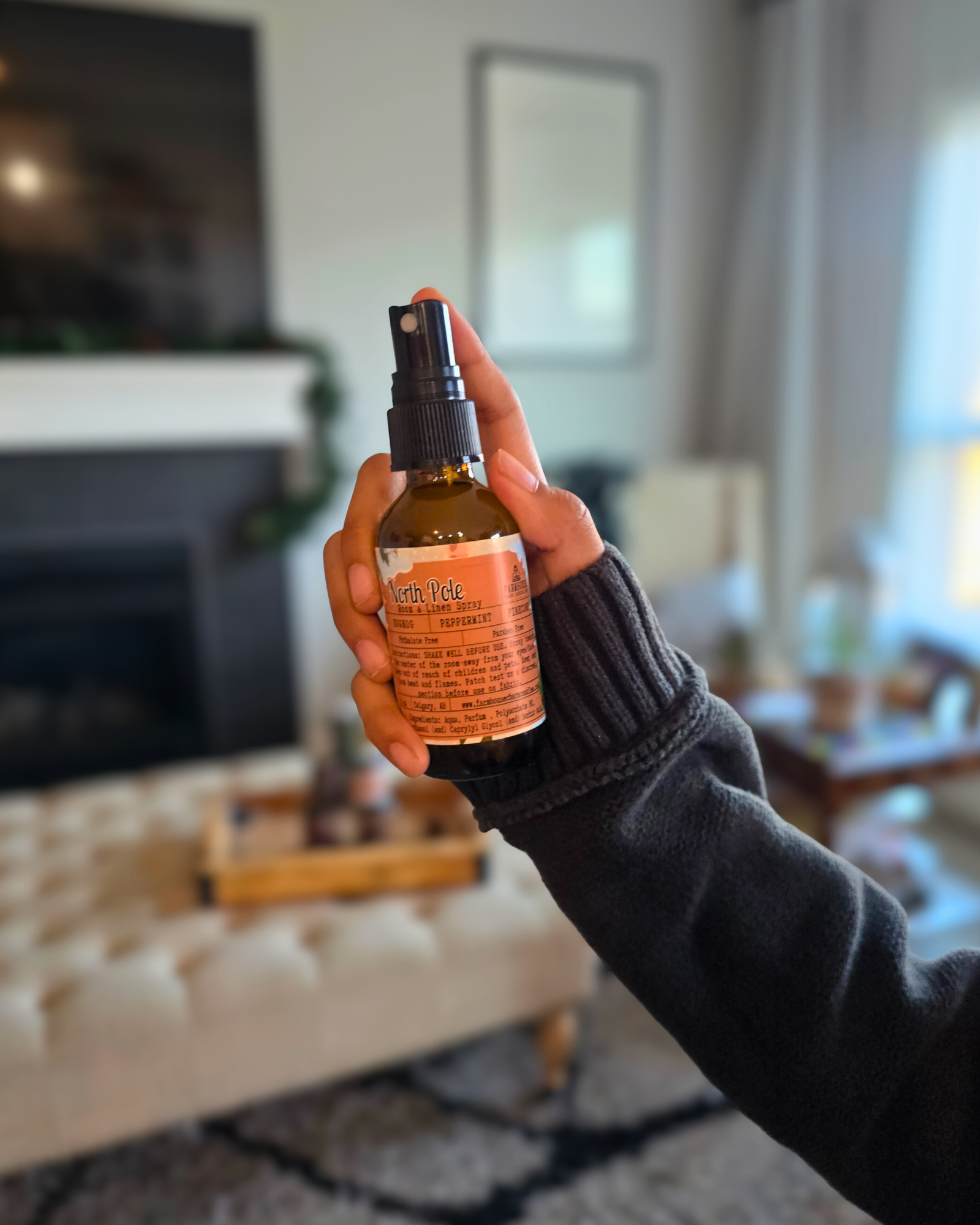 Hand holding a bottle of North Pole Room & Linen Spray  capturing the cozy Christmas scent of eggnog, peppermint, and pinecone in a living room setting.