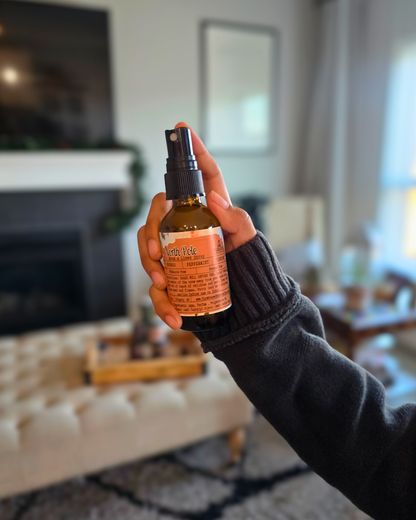 Hand holding a bottle of North Pole Room & Linen Spray  capturing the cozy Christmas scent of eggnog, peppermint, and pinecone in a living room setting.
