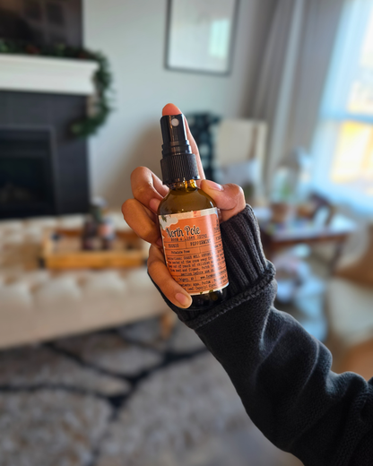 Hand holding a bottle of North Pole Room & Linen Spray  capturing the cozy Christmas scent of eggnog, peppermint, and pinecone in a living room setting.