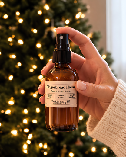 A hand holding an amber glass room and linen spray bottle labeled 'Gingerbread House' by Farmhouse Charm Candles against a blurred Christmas tree with warm lights.
