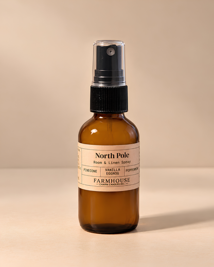 An amber glass spray bottle with a black pump dispenser and a cream-colored label reading 'North Pole' by Farmhouse Charm Candles photographed against a solid beige background.