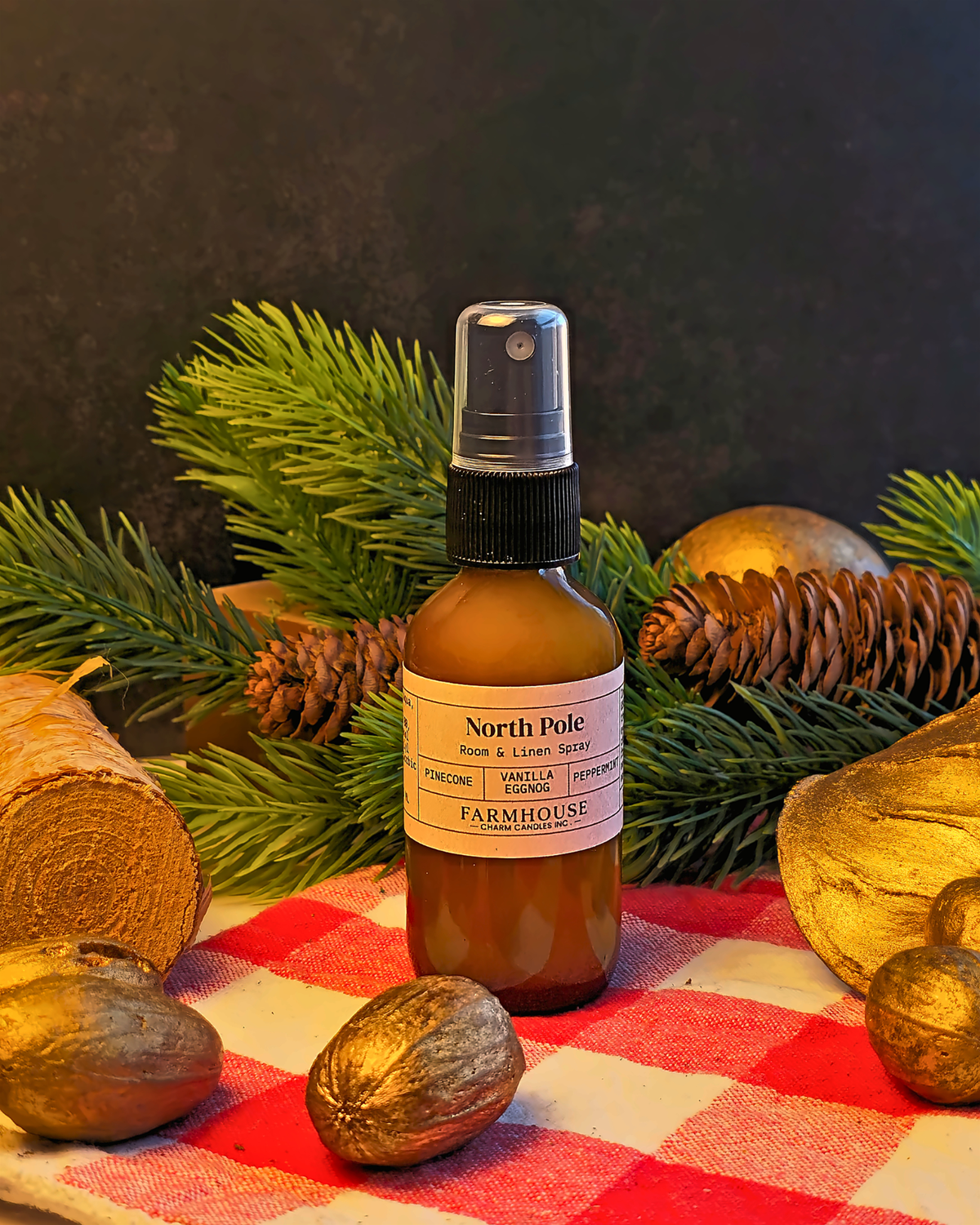 An amber glass spray bottle labeled North Pole by  Farmhouse Charm Candles set on a red and white checkered blanket with pine branches, gold pinecones, and walnuts around it.