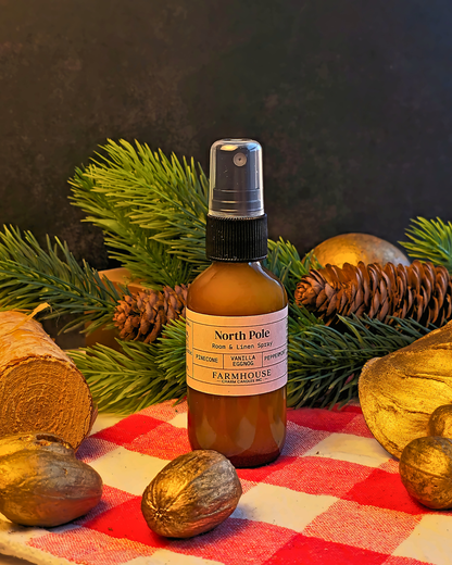 An amber glass spray bottle labeled North Pole by  Farmhouse Charm Candles set on a red and white checkered blanket with pine branches, gold pinecones, and walnuts around it.