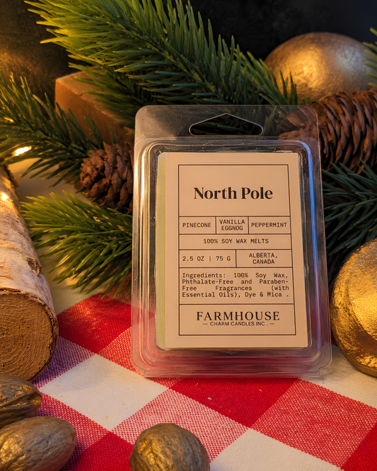 A packaged wax melt labeled 'North Pole' by Farmhouse Charm Candles is centered on a red-and-white checkered cloth, surrounded by pine branches, pinecones, and gold ornaments in a styled holiday setting.