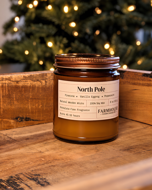 A brown amber glass jar candle with a cream label reading 'North Pole' and Farmhouse Charm Candles set on a rustic wooden surface with a blurred Christmas tree and warm lights in the background.