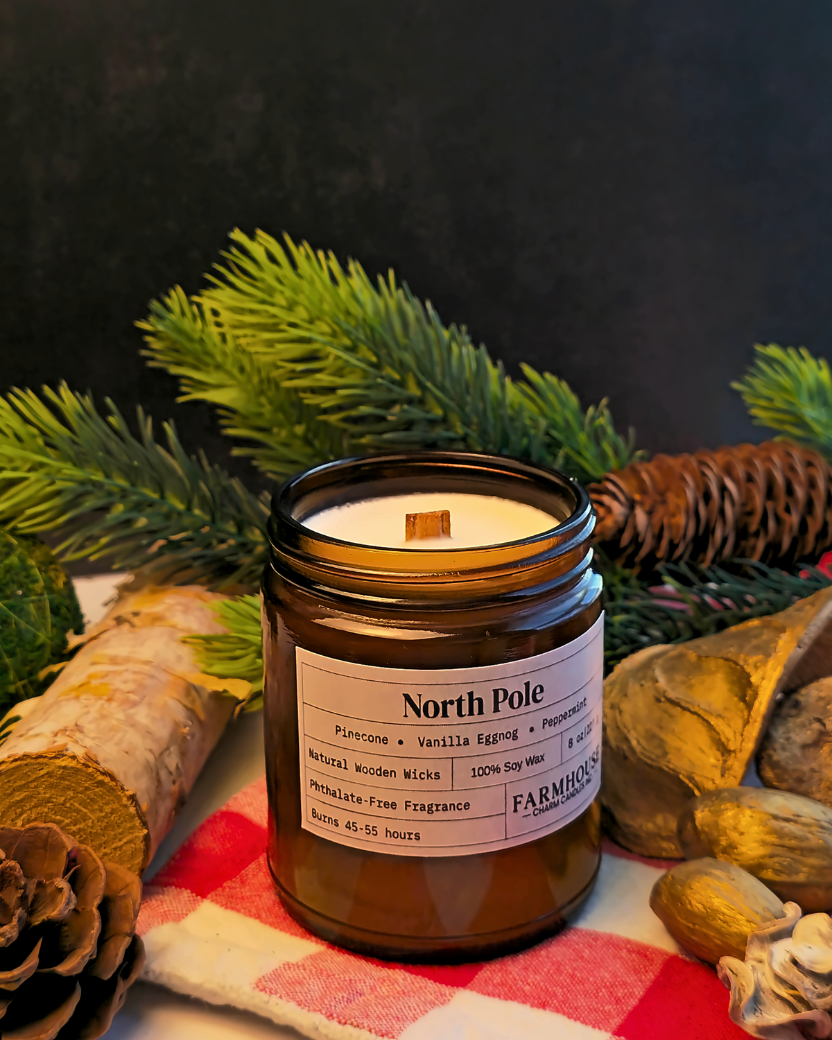 A North Pole Farmhouse Charm Candles soy wax candle in an amber glass jar, styled with pine branches, a birch log, and a red-and-white checkered cloth against a dark background.