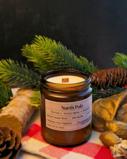 A North Pole Farmhouse Charm Candles soy wax candle in an amber glass jar, styled with pine branches, a birch log, and a red-and-white checkered cloth against a dark background.
