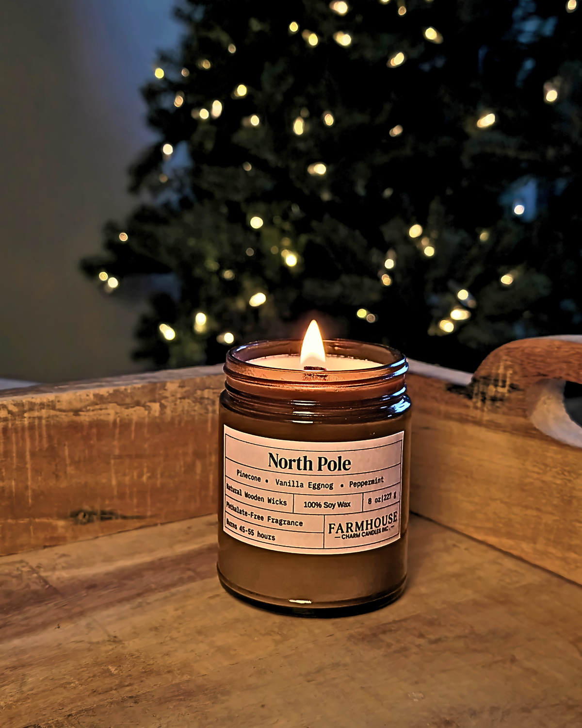 A lit amber glass jar candle labeled 'North Pole' by Farmhouse Charm Candles sits on a rustic wooden surface with a blurred Christmas tree and warm lights in the background.
