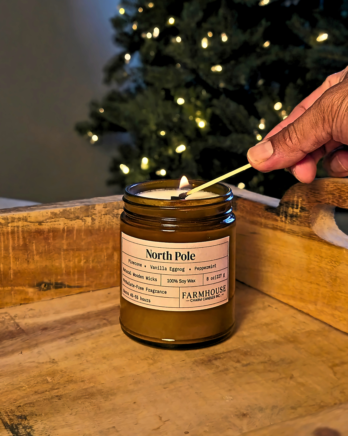 A lit candle in a brown glass jar sits on a wooden surface with a hand holding a match near the flame. A blurred Christmas tree with warm lights is in the background. North Pole by Farmhouse Charm Candles