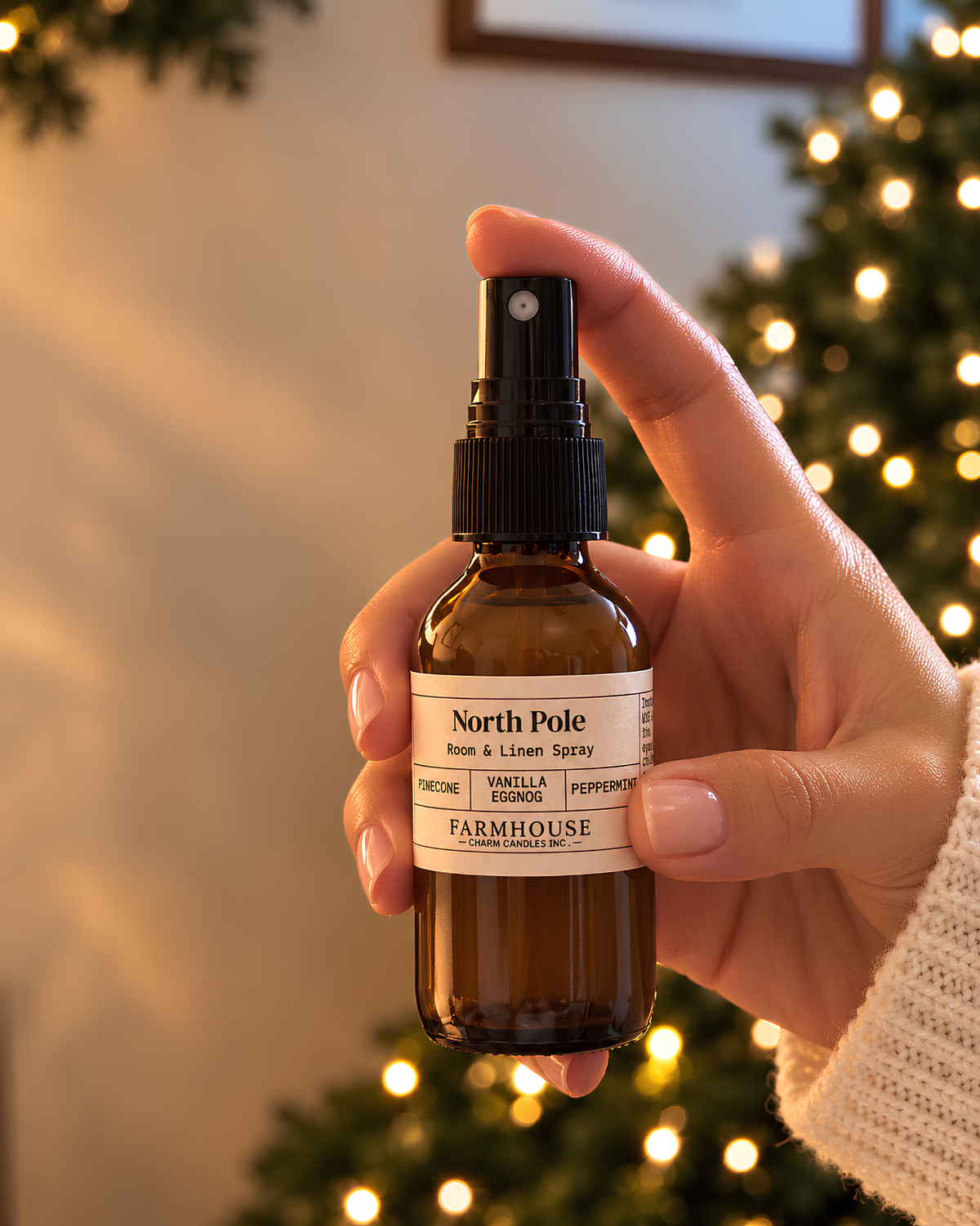A hand holding a small amber glass spray bottle labeled North Pole Room & Linen Spray by Farmhouse Charm Candles, with a blurred Christmas tree and warm lights in the background.