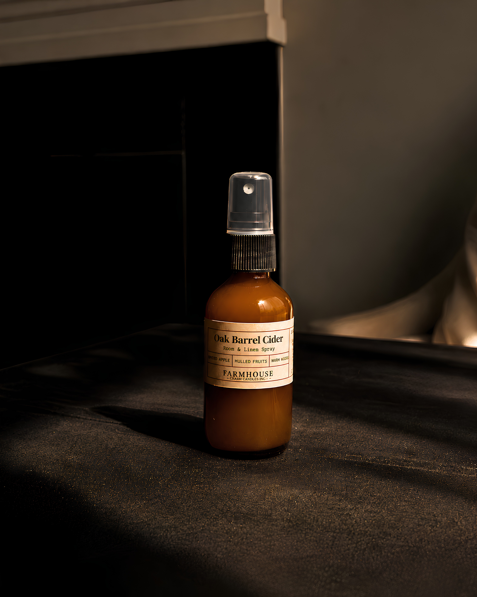 A small amber glass spray bottle labeled 'Oak Barrel Cider' by Farmhouse Charm Candles, stands on a dark tabletop in a dimly lit interior setting.