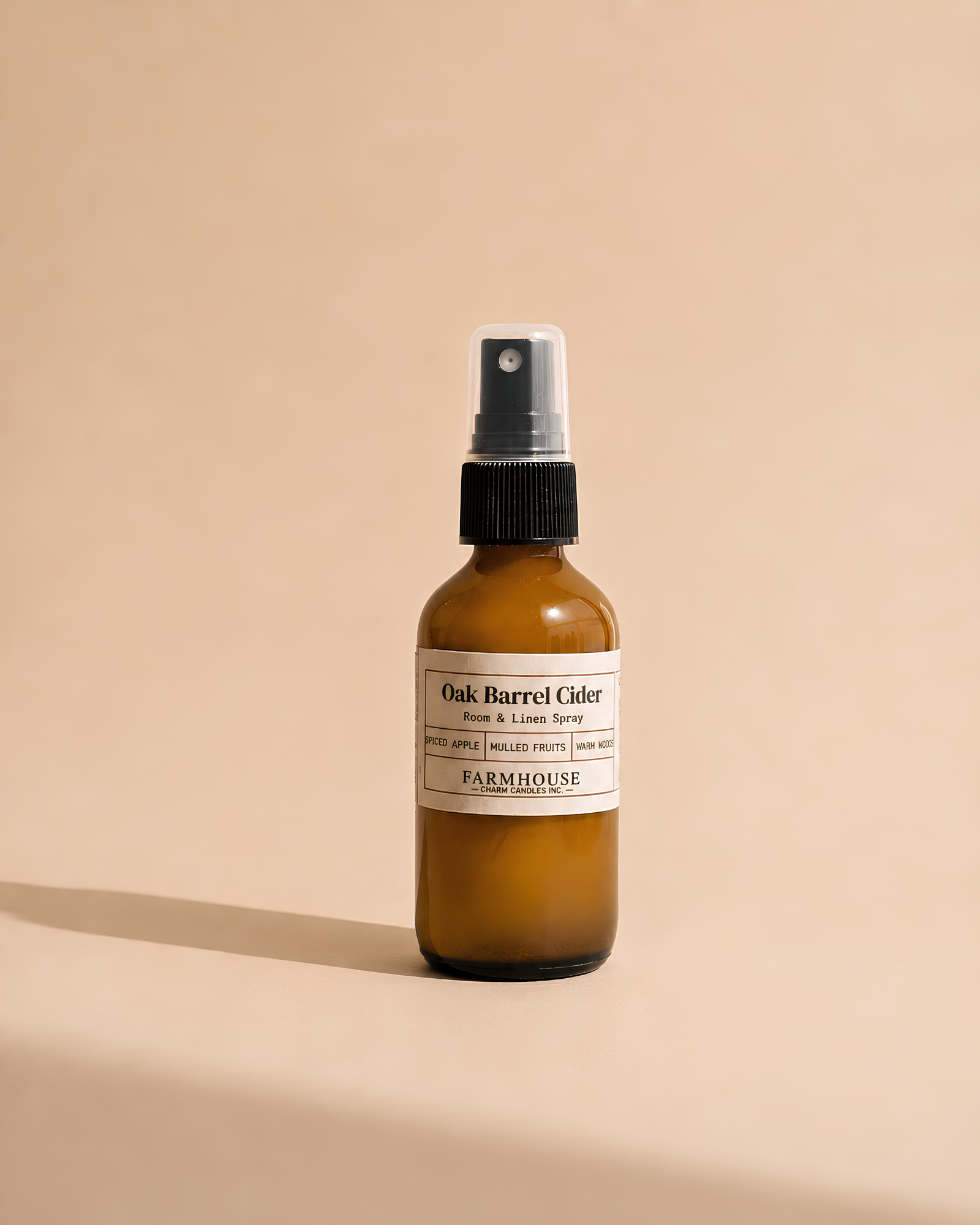 An amber glass spray bottle with a black spray cap and a cream label reading 'Oak Barrel Cider Room & Linen Spray' by Farmhouse Charm Candles, photographed on a warm beige background with a soft shadow.
