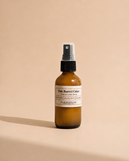 An amber glass spray bottle with a black spray cap and a cream label reading 'Oak Barrel Cider Room & Linen Spray' by Farmhouse Charm Candles, photographed on a warm beige background with a soft shadow.