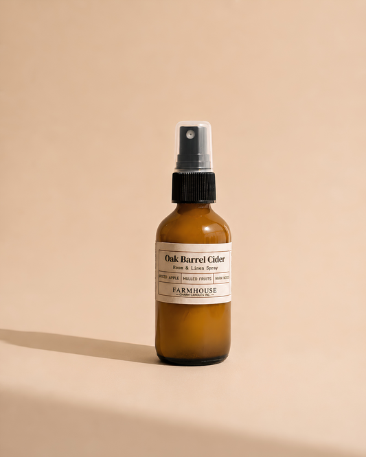 An amber glass spray bottle with a black spray cap and a cream label reading 'Oak Barrel Cider Room & Linen Spray' by Farmhouse Charm Candles, photographed on a warm beige background with a soft shadow.