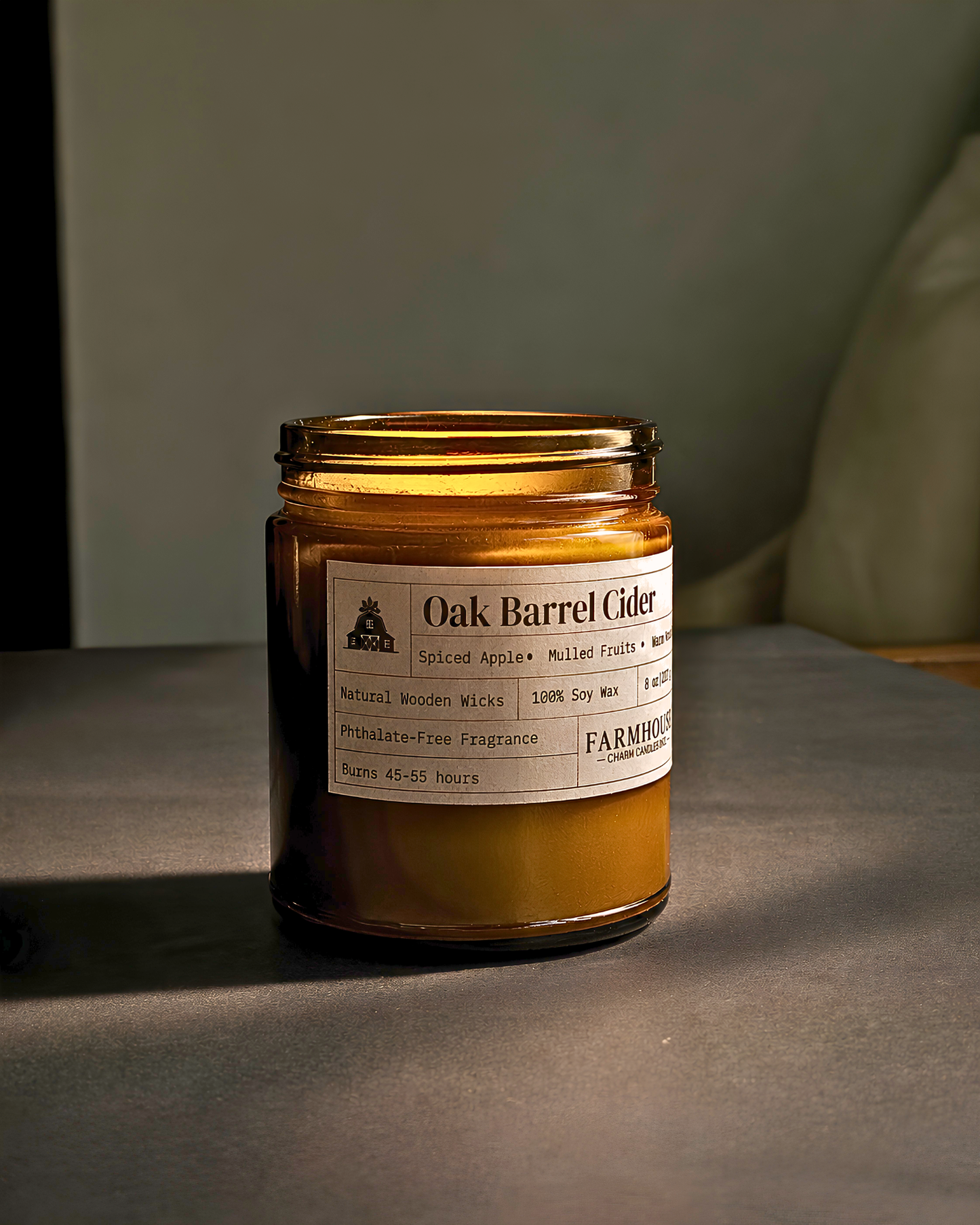 An amber glass jar candle labeled 'Oak Barrel Cider' by Farmhouse Charm Candle , set on a dark gray surface with a soft-focus background.