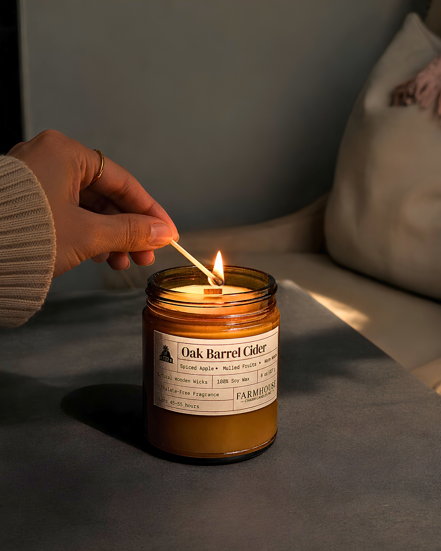 A lit amber glass jar candle on a dark surface, with a hand holding a match above the flame. The candle label reads 'Oak Barrel Cider' by Farmhouse Charm Candles
