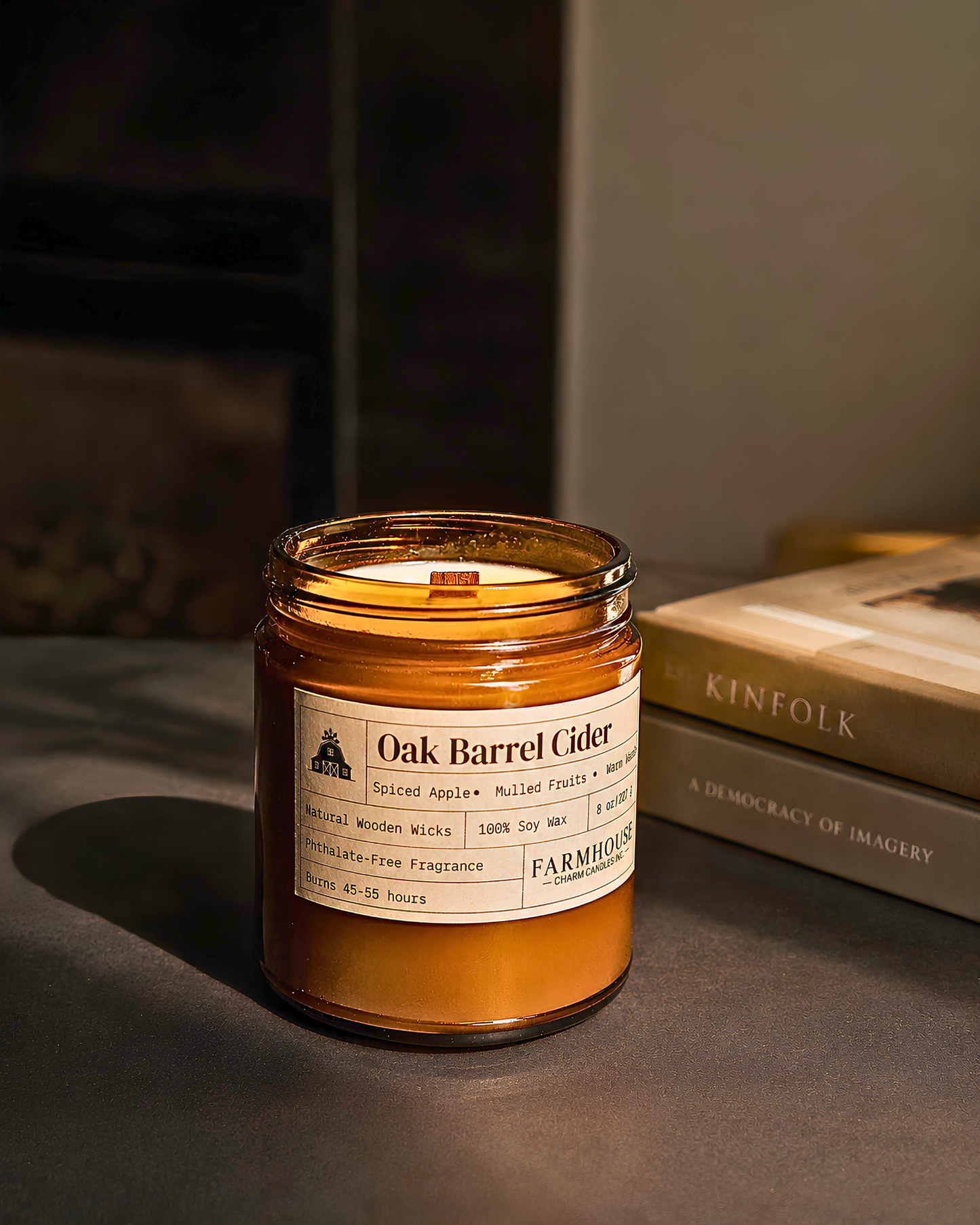 A Farmhouse Charm Candles brand 'Oak Barrel Cider' scented candle in an amber glass jar, styled on a dark surface next to a book.